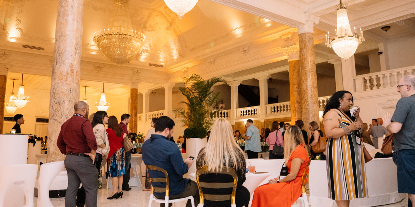 Elegant indoor event at Antiguo Casino with people mingling and seated at tables in a grand hall featuring marble columns, ornate chandeliers, and warm ambient lighting. The space includes white furniture, decorative plants, and a sophisticated architectural design with a balcony and high ceilings
