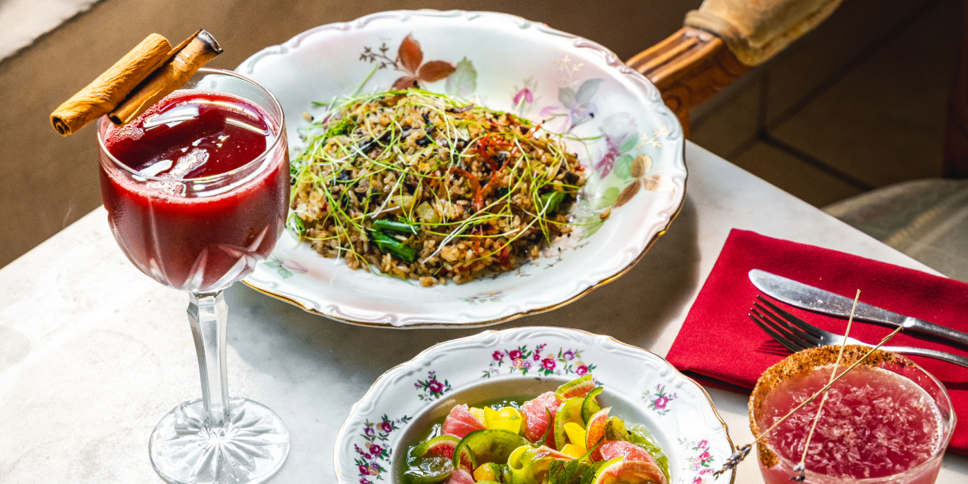 Elegant table setting with a plate of seasoned rice and vegetables topped with microgreens, a colorful salad with citrus slices in a floral-patterned dish, and two drinks—one deep red garnished with a cinnamon stick and another pink with a sugared rim—placed beside a red napkin and silverware.