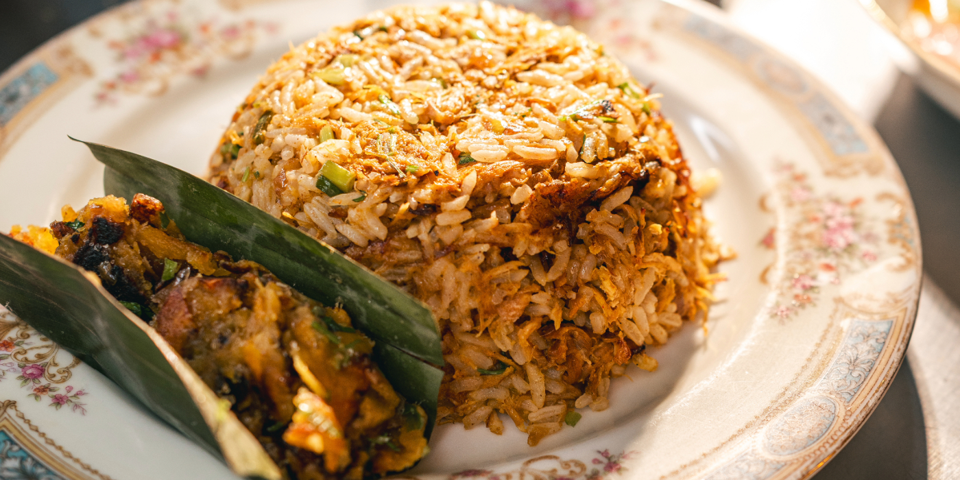 lose-up of a plate with a dome-shaped serving of seasoned rice, garnished with herbs, accompanied by a side of cooked vegetables wrapped in a green leaf, all presented on a decorative floral-patterned plate