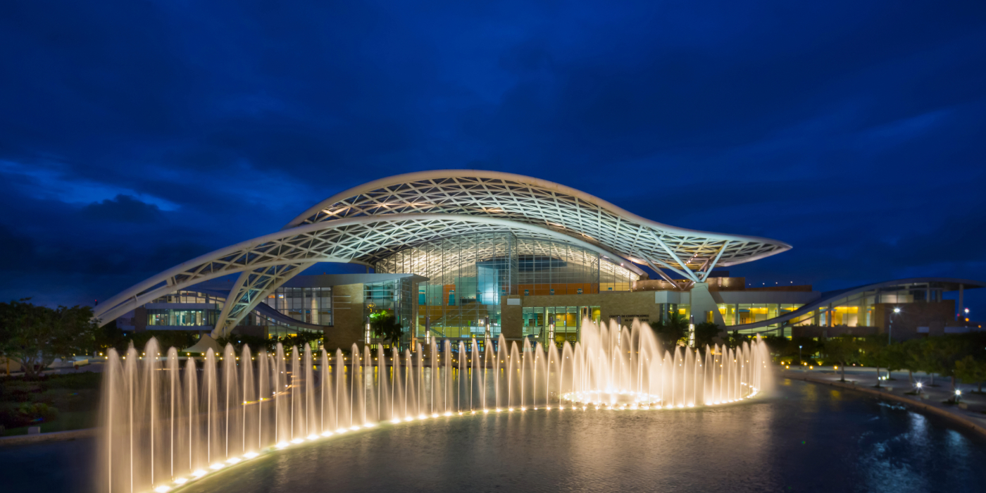 Night view of the Puerto Rico Convention Center featuring its modern, curved architectural roof illuminated with bright lights, a large circular fountain with streams of water in the foreground, and a deep blue evening sky in the background.