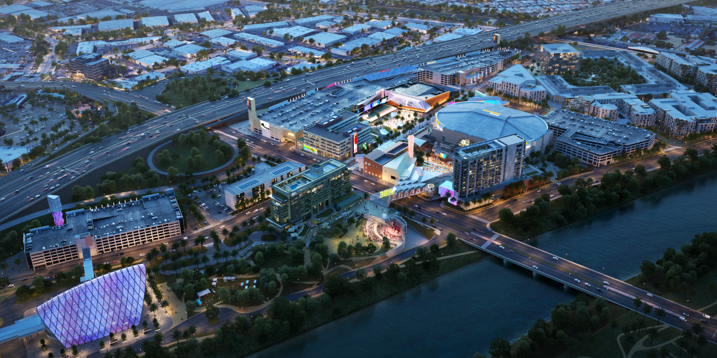 A nighttime aerial rendering of a large urban entertainment and commercial district featuring modern buildings, a brightly lit arena, parking structures, and colorful architectural lighting. A river runs along the foreground with a bridge connecting to the complex, and a network of roads and highways surrounds the development. The surrounding cityscape extends into the distance.