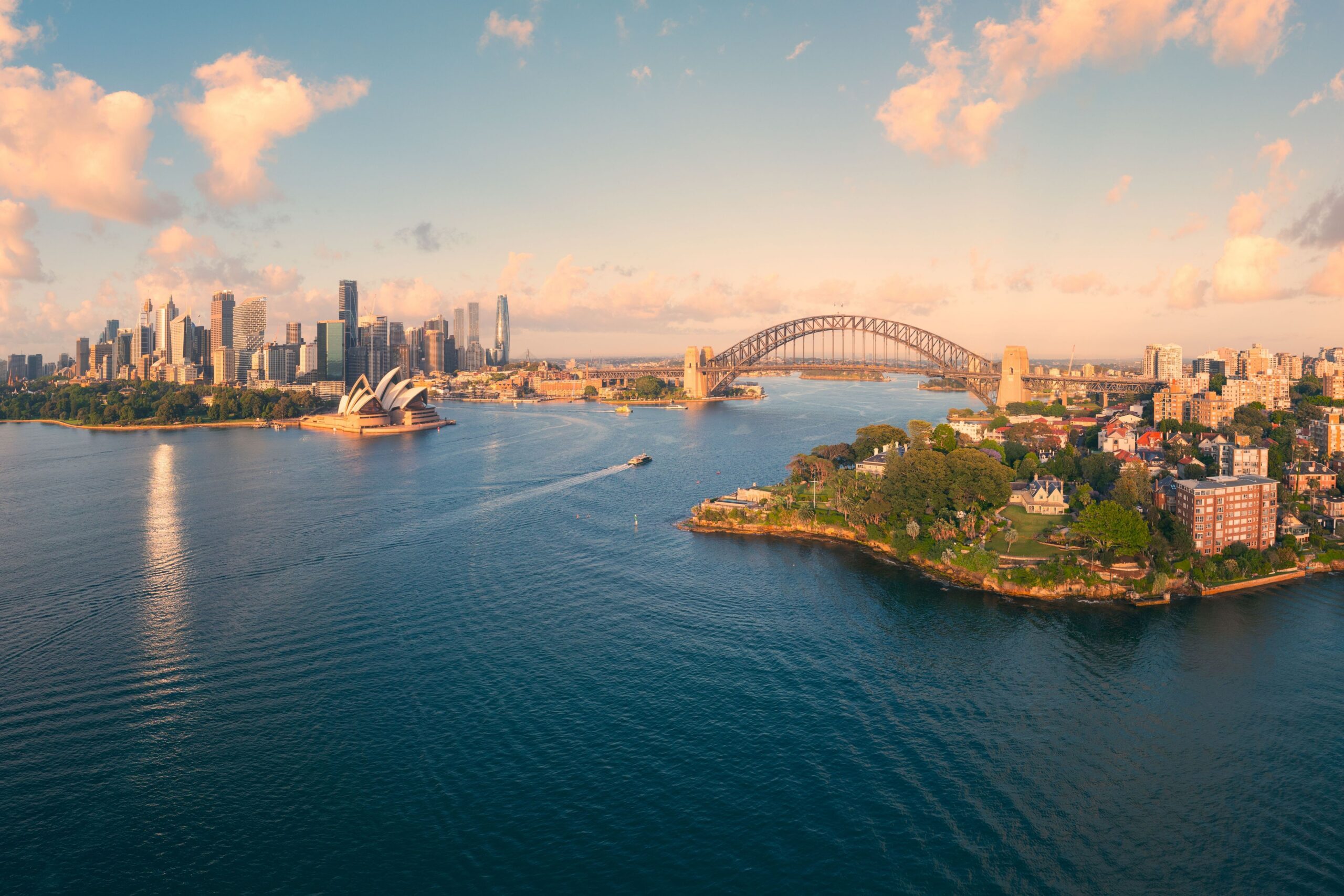 A wide aerial view of Sydney Harbour at sunset, featuring the Sydney Opera House, the Sydney Harbour Bridge, the city skyline, and boats moving across the water, with warm light reflecting off the harbour. Provide your feedback on BizChat