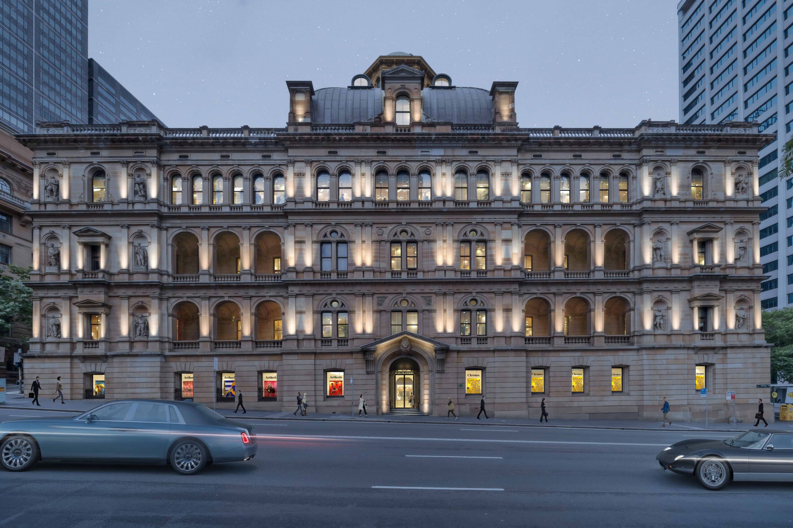 A grand, historic sandstone building illuminated at dusk, featuring arched windows, ornate architectural details, and a central arched entrance, with cars passing on the street in front and pedestrians walking along the sidewalk.
