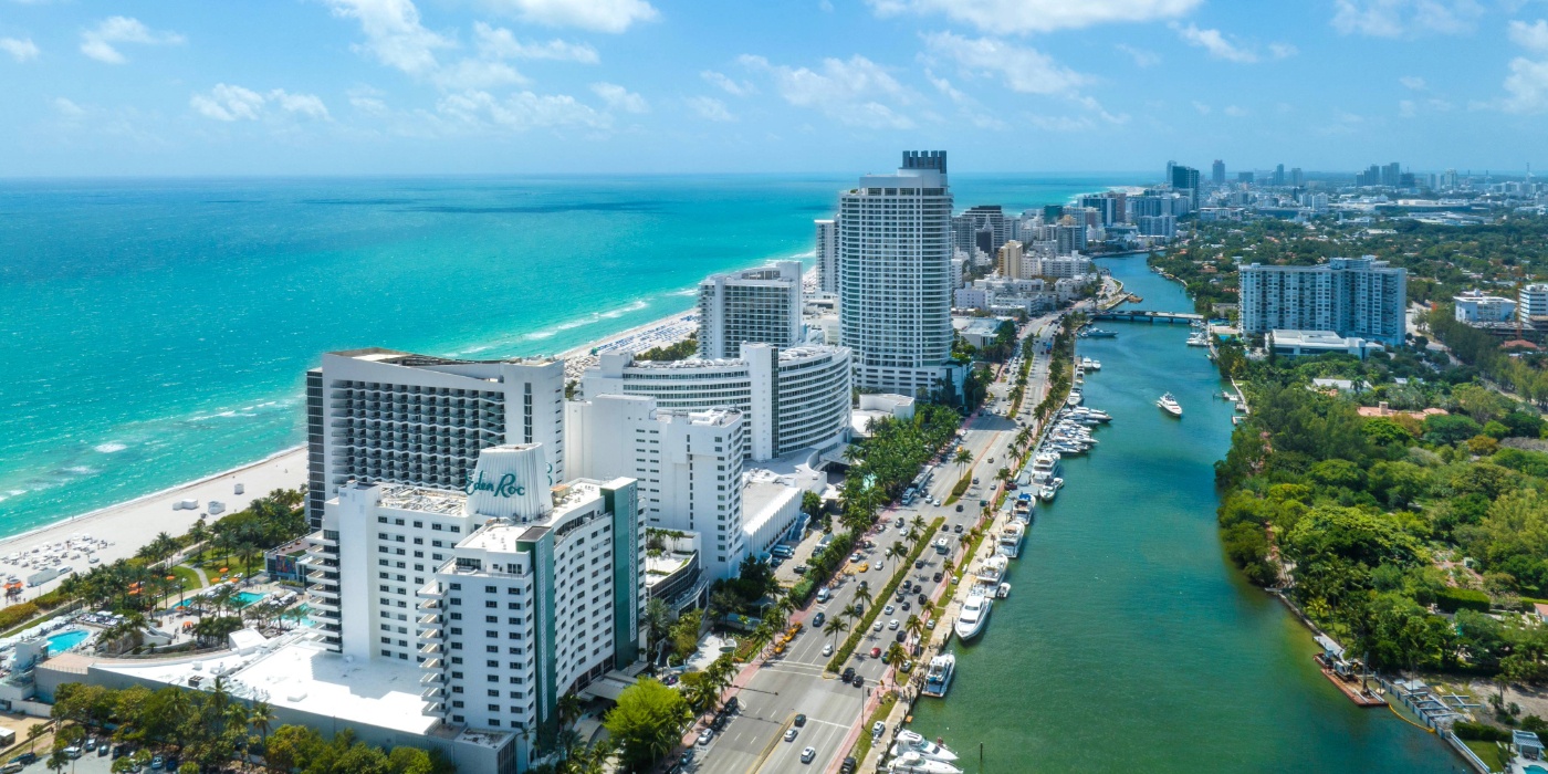 Aerial view of Miami’s Mid Beach coastline with beachfront hotels, turquoise ocean, and a canal lined with boats.
