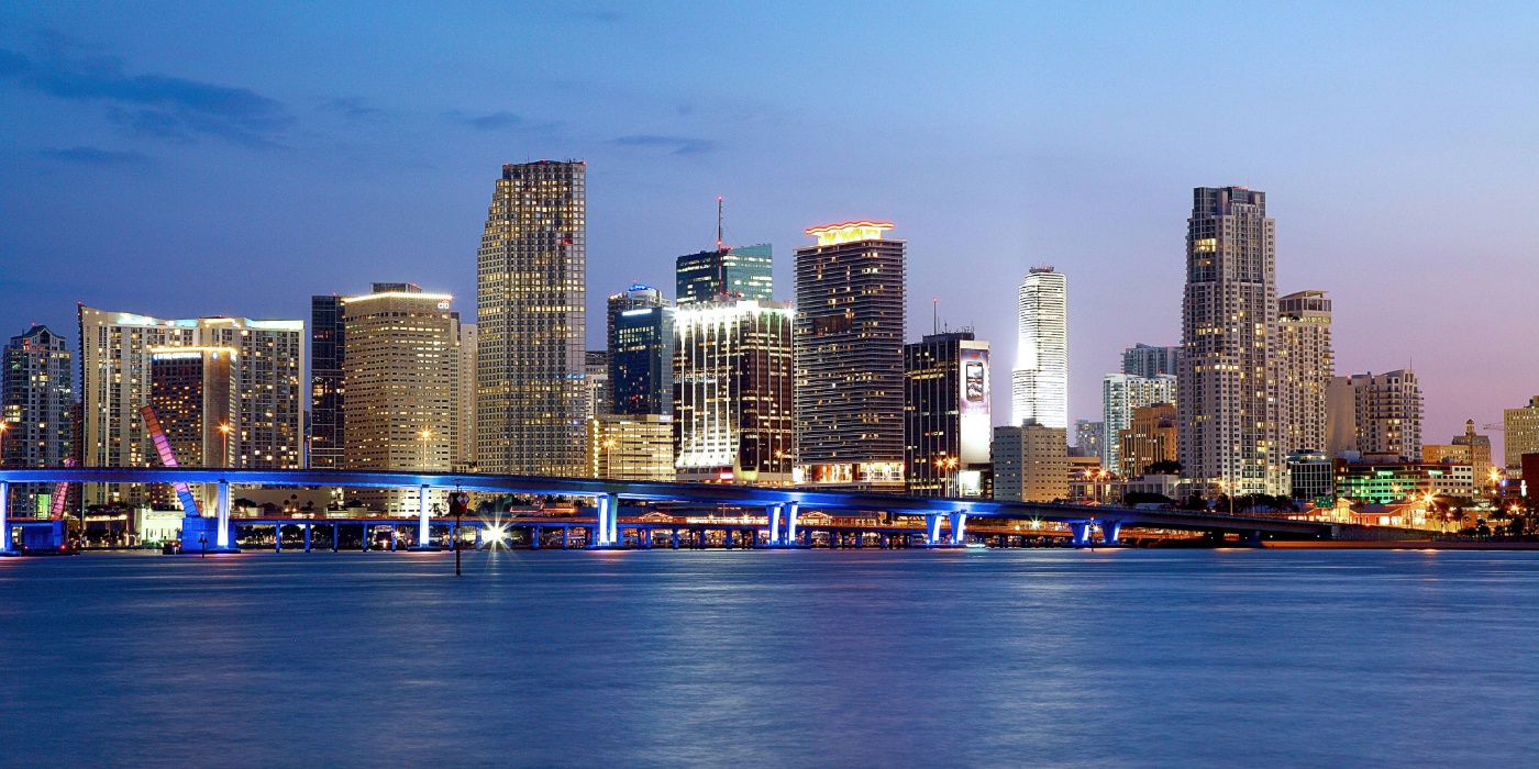 Downtown Miami skyline at dusk with illuminated skyscrapers reflected on the water.